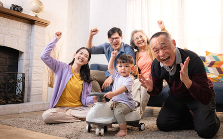Happy multi-generational Asian family enjoying quality time together at home, cheering and smiling with a toddler riding a toy car in a cozy living roomの写真素材