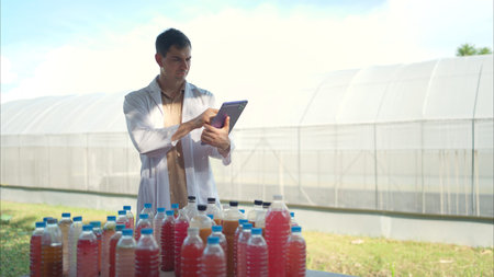 A researcher in a lab coat uses a tablet to inspect colorful liquid samples in plastic bottles, set...
