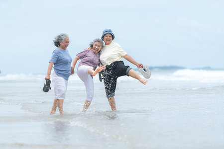 Happy senior women enjoying a playful walk on the beach, laughing and holding hands as they splash...