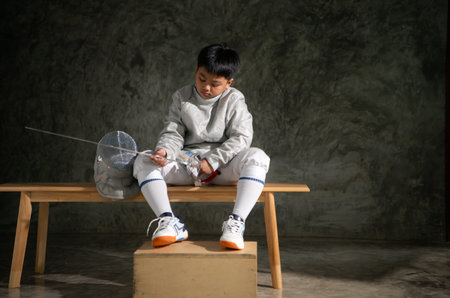 Young boy in fencing gear sitting on a bench, examining his Ã©pÃ©e sword with focus. Helmet...