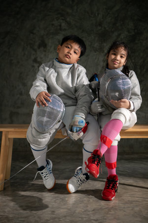 Two child in fencing gear sit on a bench, smiling at each other. They hold their masks and swords,...