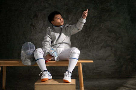 Young boy in fencing gear sitting on a bench, examining his Ã©pÃ©e sword with focus. Helmet...
