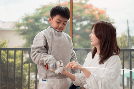 A boy in fencing gear with their mother preparing sports equipment before practicing fencing in...
