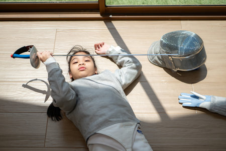 A girl in fencing gear is sitting and rest on the sidelines, exhausted from the training that just...