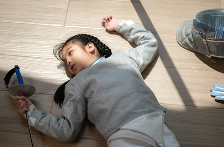 A girl in fencing gear is sitting and rest on the sidelines, exhausted from the training that just...
