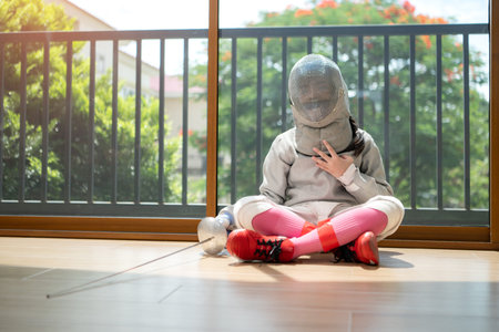 A girl in fencing gear is sitting and rest on the sidelines, exhausted from the training that just...