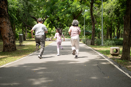 Happy Asian grandparents and granddaughter running joyfully together in a park, Active multigenerational family having fun outdoors in nature on a sunny dayの写真素材