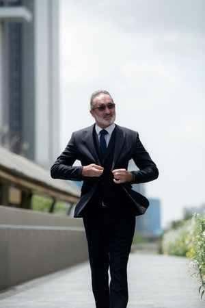 A senior businessman in a suit is walk in a hurry, with city buildings in the backgroundの写真素材