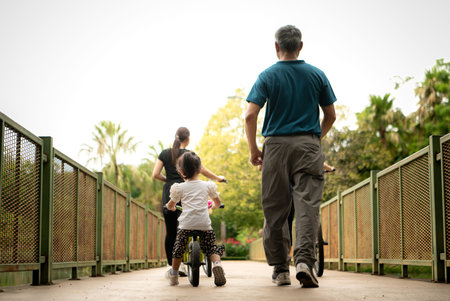 Grandfather teaching little girl to ride balance bike in park while family supports, showing love,...