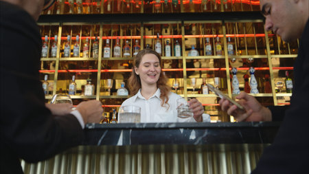 Smiling bartender serving drinks to customers at a modern bar with a stylish liquor display