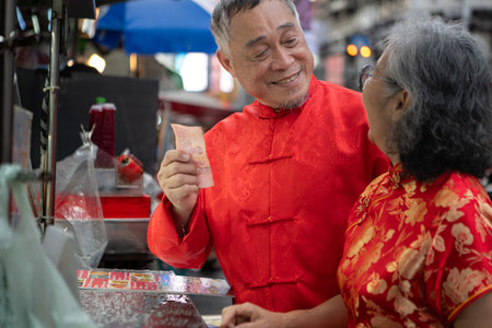 A happy senior couple strolls along a Chinatown in Bangkok, Thailand, holding hands and sightseeing together, enjoying travel and enjoying eating snacksの写真素材
