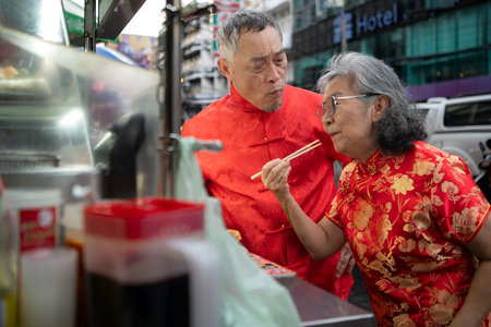 A happy senior couple strolls along a Chinatown in Bangkok, Thailand, holding hands and sightseeing together, enjoying travel and enjoying eating snacksの写真素材