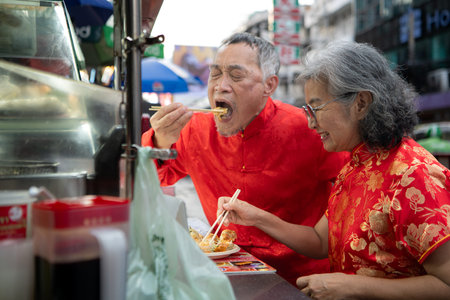 A happy senior couple strolls along a Chinatown in Bangkok, Thailand, holding hands and sightseeing together, enjoying travel and enjoying eating snacksの写真素材