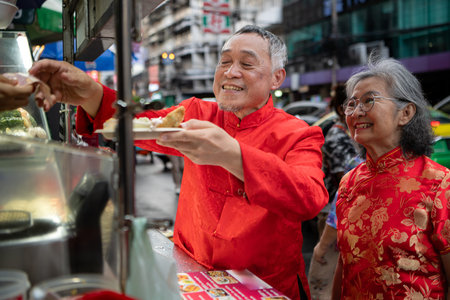 A happy senior couple strolls along a Chinatown in Bangkok, Thailand, holding hands and sightseeing together, enjoying travel and enjoying eating snacksの写真素材