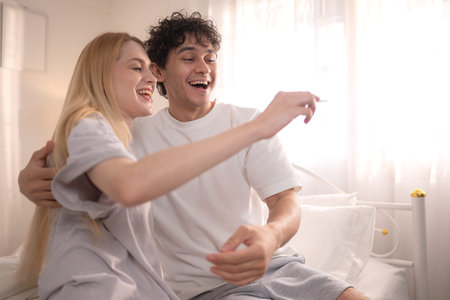 Happy couple sitting on the bed and smiling with excitement while looking at a positive pregnancy testの写真素材