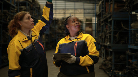 Two female engineers wearing safety gear inspecting car parts inventory in an industrial warehouse