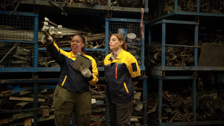 Two female engineers wearing safety gear inspecting car parts inventory in an industrial warehouse