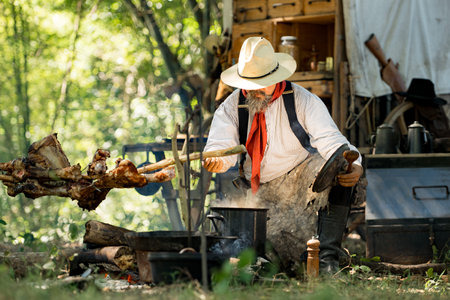 A cowboy preparing food at a rustic outdoor campfire, seasoning and stirring a steaming soup pot while roasting meat beside himの写真素材