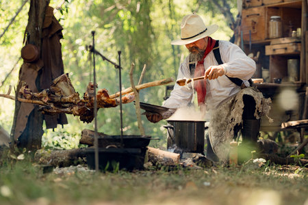A cowboy preparing food at a rustic outdoor campfire, seasoning and stirring a steaming soup pot while roasting meat beside himの写真素材