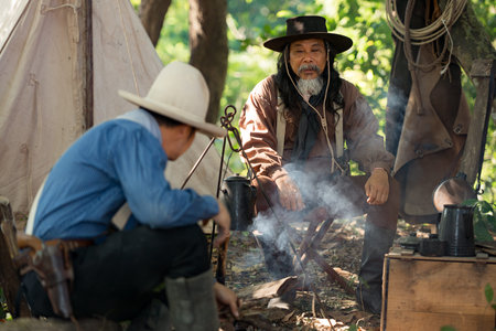 Two cowboys preparing food at a campfire, roasting meat together in a warm and rustic outdoor settingの写真素材