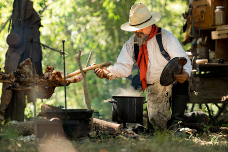 A cowboy preparing food at a rustic outdoor campfire, seasoning and stirring a steaming soup pot while roasting meat beside himの写真素材