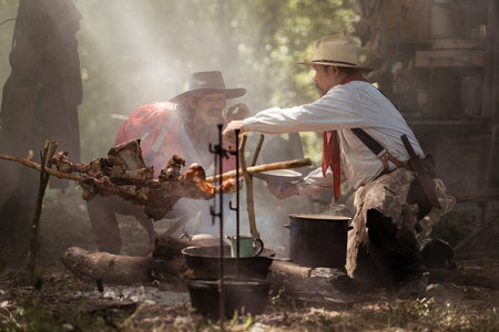 Two cowboys preparing food at a campfire, roasting meat together in a warm and rustic outdoor settingの写真素材