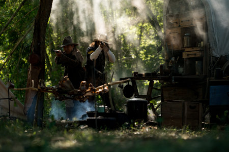 Two cowboys aiming rifles in a forest camp, with sunlight streaming through the smoke and a chuck wagon in the backgroundの写真素材
