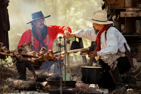 Two cowboys preparing food at a campfire, roasting meat together in a warm and rustic outdoor settingの写真素材