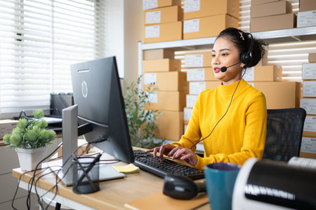 Customer service representative wearing headset working at computer in warehouse office, managing online orders and supportの写真素材