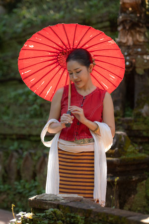 Asian woman wearing traditional Thai dress holding a red umbrella while walking along a garden path surrounded by natureの写真素材