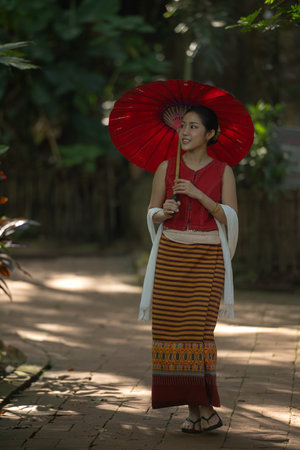 Asian woman wearing traditional Thai dress holding a red umbrella while walking along a garden path surrounded by natureの写真素材