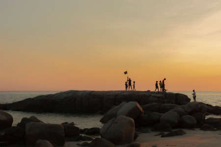 The stones on beach in evening have the background of the sea and the evening sun. There are people standing on rocks and flagsの写真素材