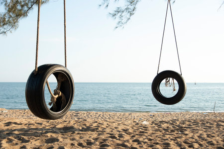 Tire swings hang from trees on the sandy beach. With a background of blue sea and sky with coludsの写真素材