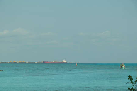 A small blue fishing boat was sailing in the middle of the sea, heading for the shore. There is a sky and a large cargo ship as a backdrop.の写真素材