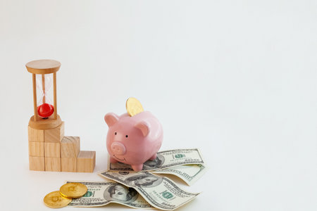 Pig shaped piggy bank with bitcoin coins, hourglass, growing graph wood cubes and us dollars on isolated white backgroundの写真素材