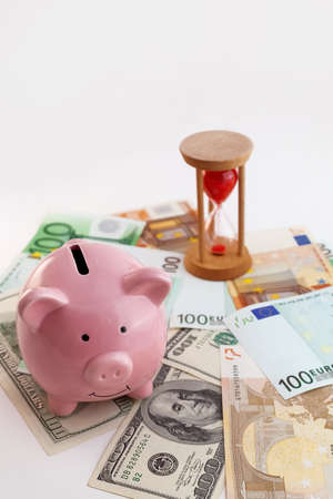 Pig shaped piggy bank, hourglass, growing graph wood cubes, euro banknotes and us dollars on isolated white backgroundの写真素材
