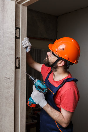 a worker with a drill in an orange helmet installs a door in the houseの写真素材