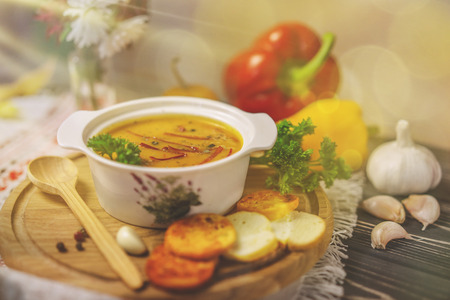 Pumpkin soup in white porcelain pot, toasts, vegetables and flowers standing on a table with nice wooden texture. Sunny day and tasty lunch. Vintage effect.の写真素材