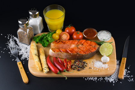 Grilled salmon steak with sauces, spices, vegetables, parsley and grissini breads on a wooden cutting board on a black background.の写真素材