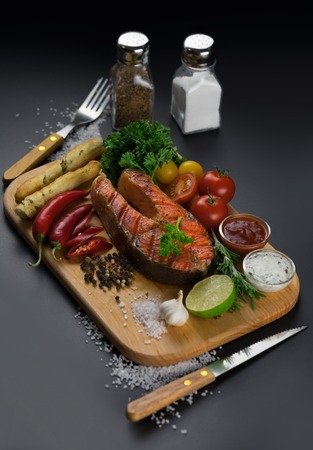 Grilled salmon steak with vegetables, sause and spices on a wooden cutting board on a black background.の写真素材