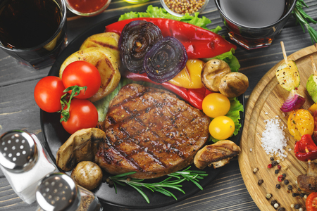 Close up of a grilled tasty beef steak with grilled potatoe, paprika, onions, mushrooms and vegetable skewers on a dark wooden background.の写真素材