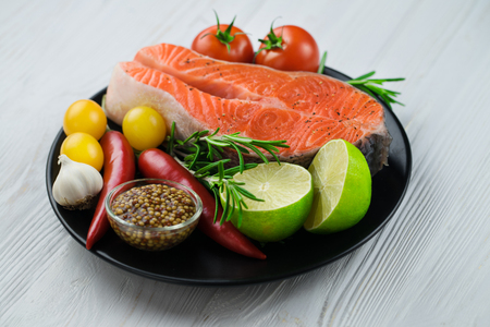Close up of two raw fresh salmon steaks on a black porcelain plate with vegetables, lime, herbs and mustard on white wooden background.の写真素材