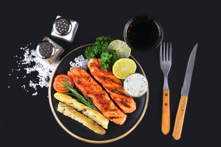 Top view of griled salmon steaks with lime, herbs, sauce and grissini bread sticks on black background.の写真素材