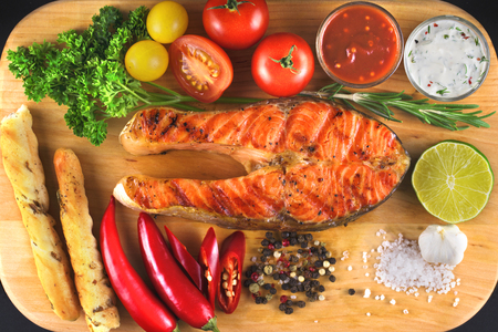 Close up top view of grilled salmon steak with vegetables, greens, sauce and grissini bread sticks on wooden cutting board.の写真素材