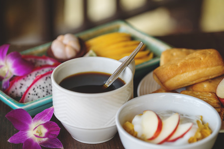 Close up of continental breakfast, corn flakes with yoghurt, coffee with  buns and cookies, and mixed tropical fruits plate on dark wooden background.の写真素材