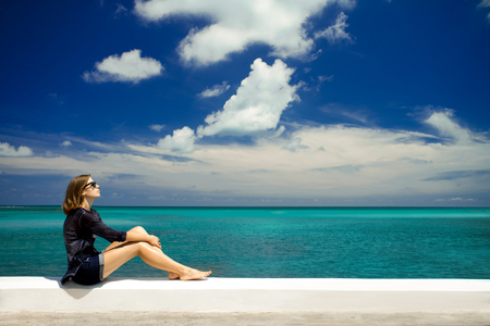 Young attractive female tourist with bob cut sitting at the embankment with the beautiful turquoise ocean water and bright blue sky on the background. Copy space for your text.の写真素材