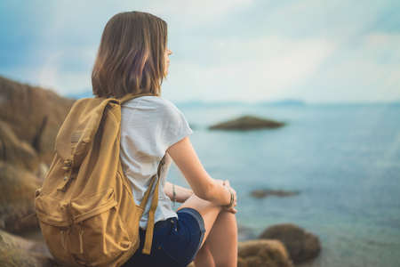 Young female hipster traveller with a backpack sitting on the rock and looking at the sea waiting for the sunrise.の写真素材