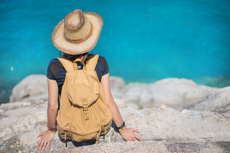 Young european female tourist with a backpack and bamboo cowboy style hat sitting on a cliff and looking at the sea. Copy space for your text.の写真素材