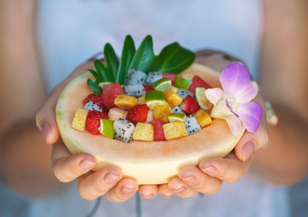 Close up of female hands holding melon half with various tropical fruits salad. Watermelon, mango, dragon fruit, lime, banana and strawberry decorated with orchid flower.の写真素材