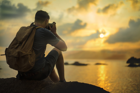 Young european male tourist with a backpack sitting at the cliff by the sea and taking photo of beautiful sunset by his retro camera. Mock up, copy space for your text.の写真素材